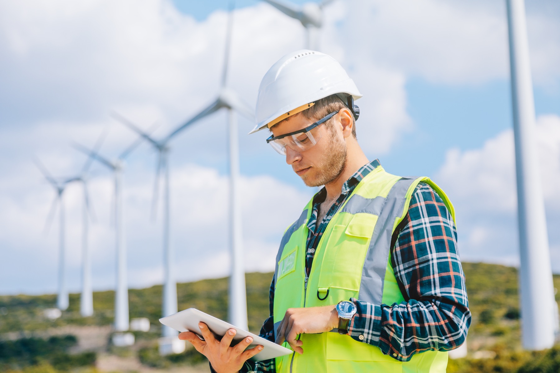 Portrait of young caucasian technician engineer man wearing white worker headwear standing, working, checking farm field system and looking up verify at wind turbine while using digital tablet pc computer for renewable energy plan by wind turbine power generation station in rural landscape. xxxl size